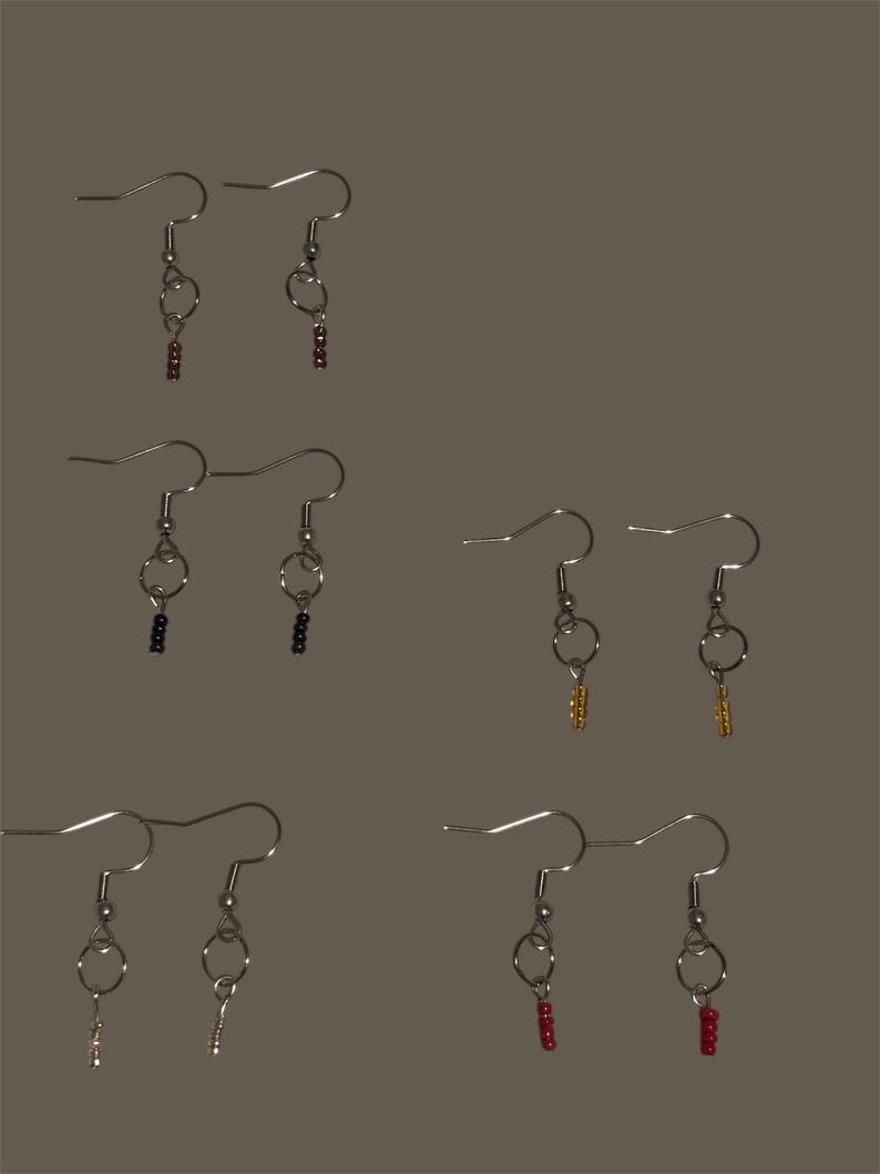 May include: Six pairs of handmade earrings with silver-colored hooks and beaded dangles in various colors, including red, black, gold, and silver. The earrings are displayed against a neutral background, showcasing their delicate design and craftsmanship.