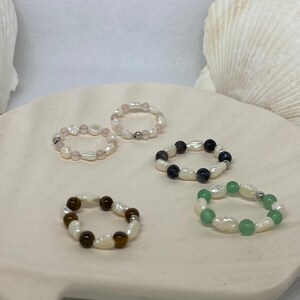 May include: Five beaded rings with different gemstone and pearl combinations. The rings feature white pearls paired with rose quartz, black and blue stones, brown tiger's eye, and green stones. The rings are displayed on a white, seashell-shaped dish.