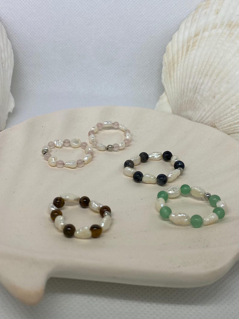 May include: Five beaded rings with different gemstone and pearl combinations. The rings feature white pearls paired with rose quartz, black and blue stones, brown tiger's eye, and green stones. The rings are displayed on a white, seashell-shaped dish.