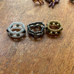 May include: A collection of beaded rings in various designs and colors. The rings include white, black, and gold beads, with some featuring multiple layers. The rings are displayed on a wooden surface, showcasing the handmade jewelry.