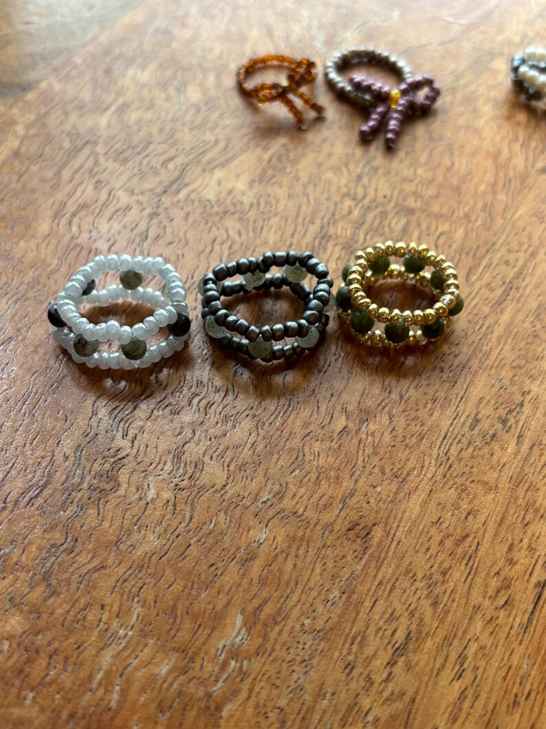 May include: A collection of beaded rings in various designs and colors. The rings include white, black, and gold beads, with some featuring multiple layers. The rings are displayed on a wooden surface, showcasing the handmade jewelry.