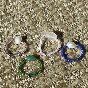 May include: Four beaded rings in various colors. One ring is green with a central green stone, another is pink with a central pink stone, a third is blue with a white pearl, and the last is pink with a white pearl.