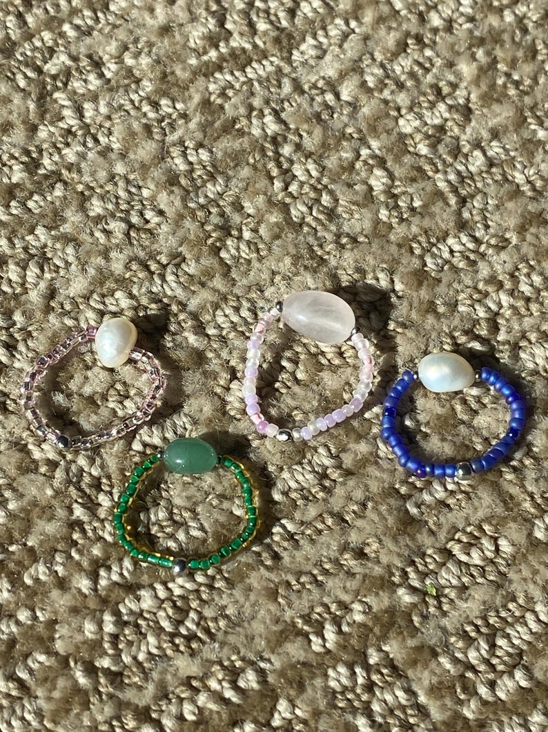 May include: Four beaded rings in various colors. One ring is green with a central green stone, another is pink with a central pink stone, a third is blue with a white pearl, and the last is pink with a white pearl.