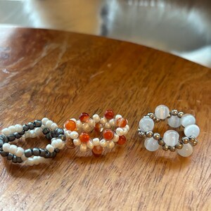 May include: Three beaded rings in a close-up shot. One ring features white and dark gray beads, another has orange and white beads, and the third has white and gold beads. The rings are on a wooden surface.