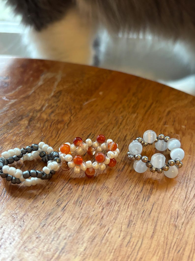 May include: Three beaded rings in a close-up shot. One ring features white and dark gray beads, another has orange and white beads, and the third has white and gold beads. The rings are on a wooden surface.