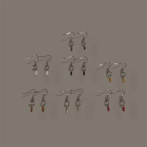 May include: A collection of handmade earrings with silver-tone hooks and various colored beaded accents. The earrings feature a key-like design, with beads in colors like white, black, gold, red, and brown. The earrings are displayed against a neutral gray background.