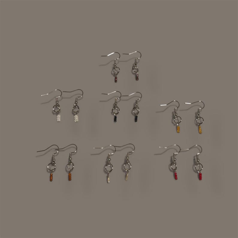 May include: A collection of handmade earrings with silver-tone hooks and various colored beaded accents. The earrings feature a key-like design, with beads in colors like white, black, gold, red, and brown. The earrings are displayed against a neutral gray background.