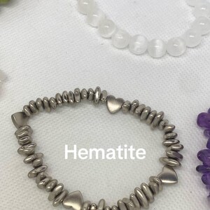 May include: A hematite bracelet with heart-shaped accents. The bracelet is silver-colored and features small, rounded beads. The word "Hematite" is printed in white on the image. Other bracelets with white and green beads are also visible.