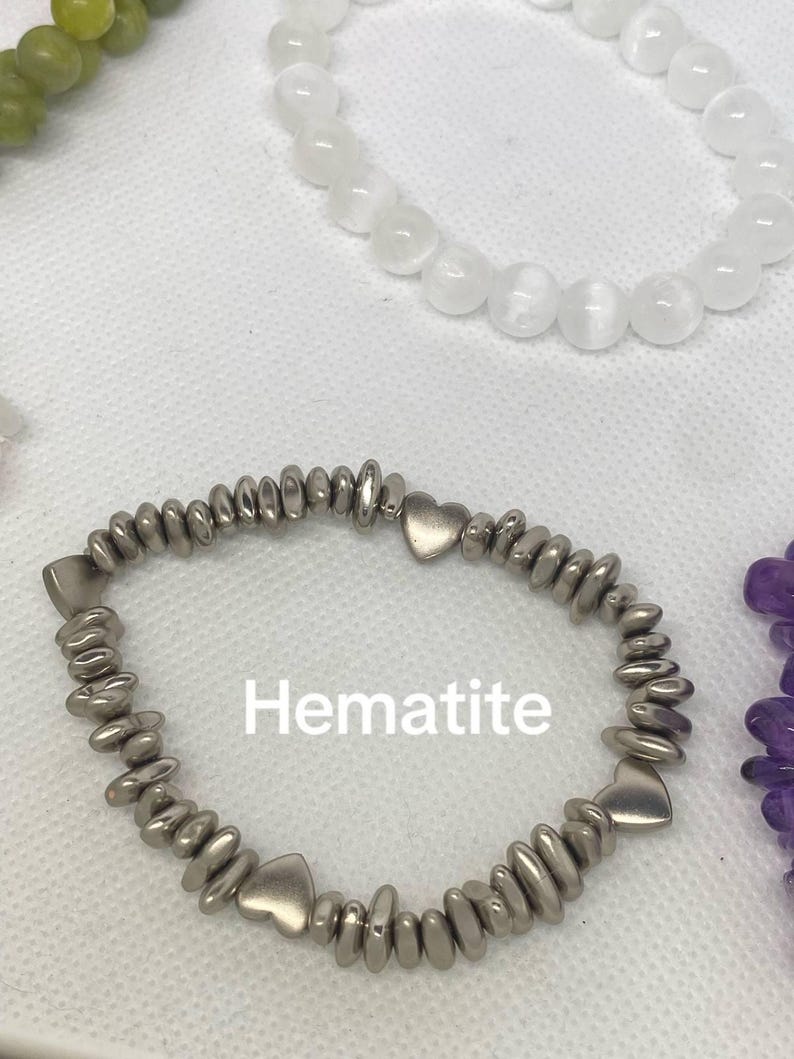 May include: A hematite bracelet with heart-shaped accents. The bracelet is silver-colored and features small, rounded beads. The word "Hematite" is printed in white on the image. Other bracelets with white and green beads are also visible.