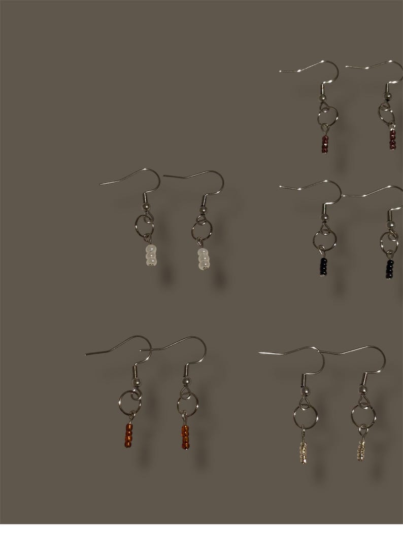 May include: A collection of handmade earrings with silver-tone hooks and circular accents. The earrings feature small, colorful beads in various shades, including white, red, black, and gold. The earrings are displayed against a neutral background.
