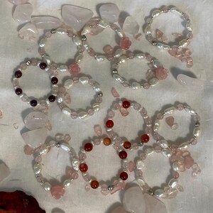 May include: A collection of beaded bracelets featuring freshwater pearls, rose quartz, and other gemstones. Some bracelets have small rose-shaped accents. The bracelets are arranged on a white surface with scattered rose quartz stones.