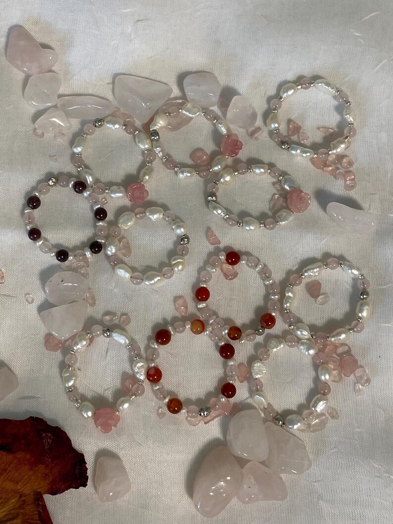 May include: A collection of beaded bracelets featuring freshwater pearls, rose quartz, and other gemstones. Some bracelets have small rose-shaped accents. The bracelets are arranged on a white surface with scattered rose quartz stones.