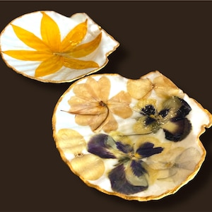 May include: Two decorative seashell-shaped dishes. One dish features a yellow flower design, while the other showcases a variety of pressed flowers in shades of yellow, purple, and white. Both have gold-painted edges.