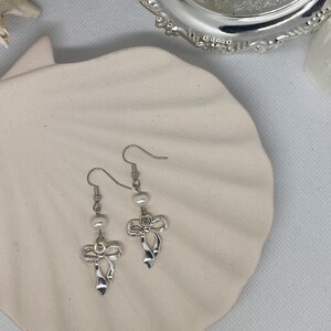 May include: A pair of silver-toned bow earrings with small, dangling pearls. The earrings are displayed on a white, seashell-shaped dish, with a silver decorative tray in the background. The earrings have a delicate, feminine design.