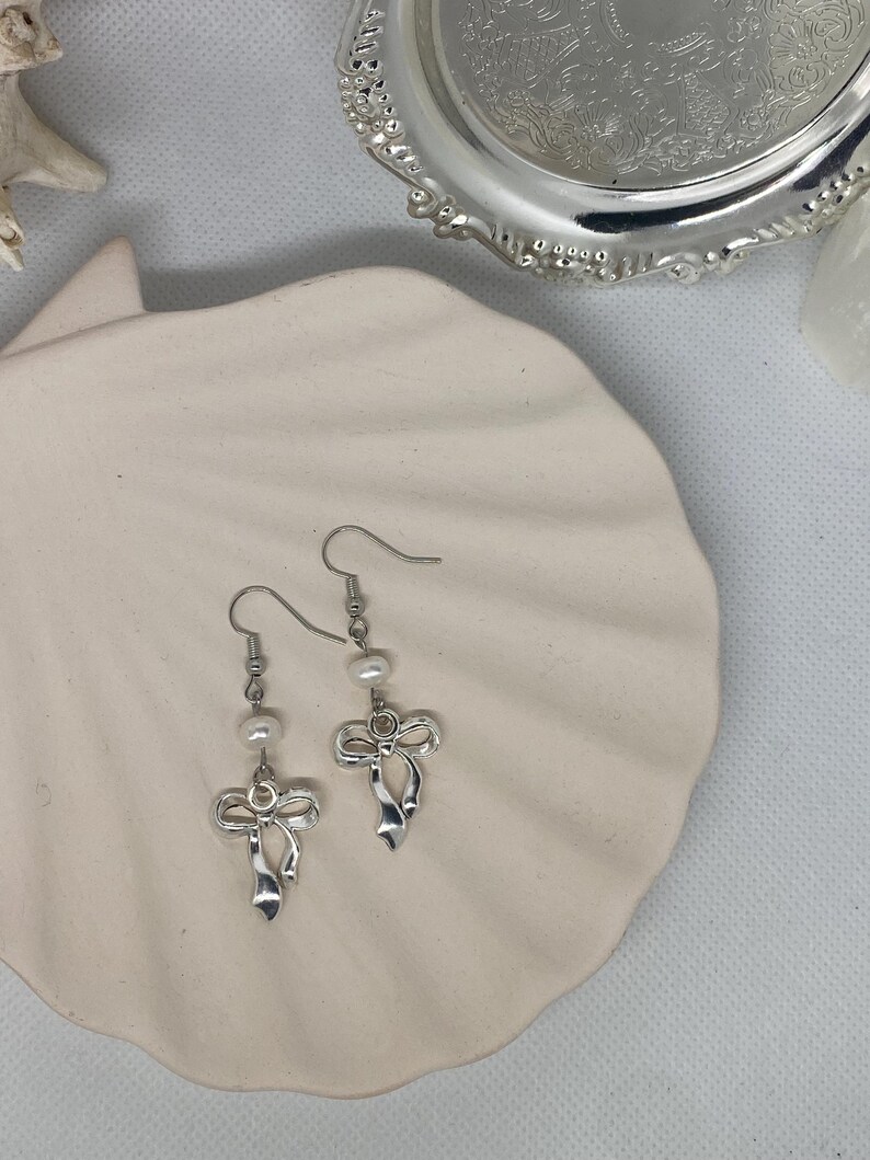 May include: A pair of silver-toned bow earrings with small, dangling pearls. The earrings are displayed on a white, seashell-shaped dish, with a silver decorative tray in the background. The earrings have a delicate, feminine design.