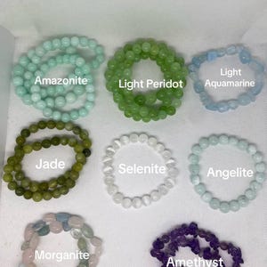 May include: An assortment of beaded bracelets in various colors and gemstones, including Amazonite, Light Peridot, Light Aquamarine, Jade, Selenite, Angelite, Morganite, and Amethyst. Each bracelet is displayed on a white surface.