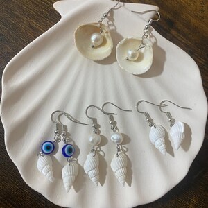 May include: A collection of handmade earrings displayed on a white seashell-shaped dish. The earrings feature natural seashells, pearls, and silver-colored hooks. Some earrings include blue evil eye charms.