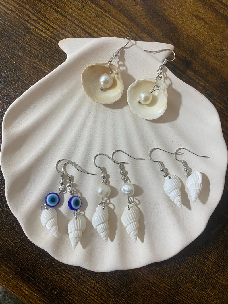 May include: A collection of handmade earrings displayed on a white seashell-shaped dish. The earrings feature natural seashells, pearls, and silver-colored hooks. Some earrings include blue evil eye charms.