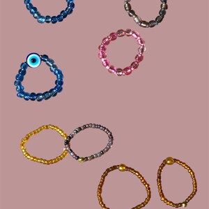 May include: A collection of beaded rings in various colors. The rings include blue with an evil eye bead, pink, gold, brown, and a ring with a mix of colors. The rings are made of small, round beads.