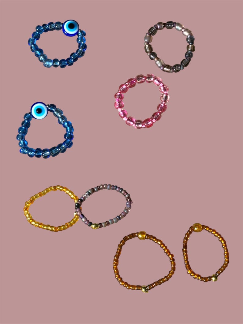May include: A collection of beaded rings in various colors. The rings include blue with an evil eye bead, pink, gold, brown, and a ring with a mix of colors. The rings are made of small, round beads.