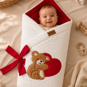 Può includere: Una coperta avvolgente per neonati bianca e testurizzata con fodera interna rossa, un grande fiocco rosso sul lato e un'applicazione di un orsacchiotto marrone che abbraccia un cuore rosso. Una piccola etichetta marrone con il testo bebella è attaccata.