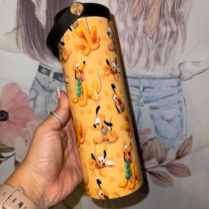 May include: A yellow tumbler with a repeating pattern of Pluto, the Disney cartoon dog. The tumbler has a black lid with a handle and a gold-coloured button. The background is a blurred image of a person.