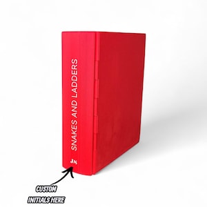 May include: A red book with the title "Snakes and Ladders" printed vertically on the spine. The book has a solid red cover and a custom initials area at the bottom. The book is standing upright against a white background.