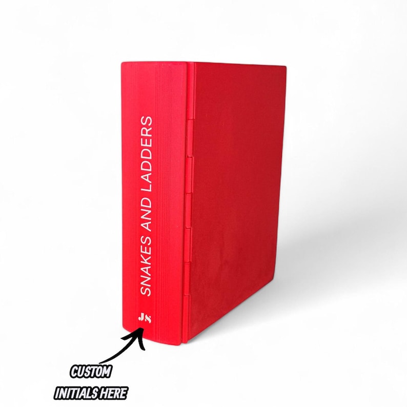 May include: A red book with the title "Snakes and Ladders" printed vertically on the spine. The book has a solid red cover and a custom initials area at the bottom. The book is standing upright against a white background.