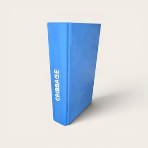 May include: A blue cribbage board with the word "CRIBBAGE" printed vertically in white. The board is rectangular and appears to be made of a solid material. The board is standing upright against a light background.