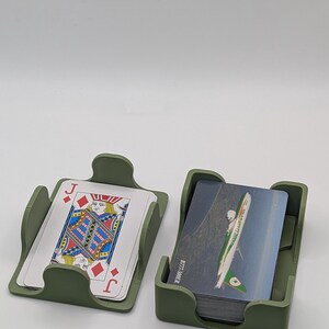 May include: Two olive green card holders, each containing playing cards. One holder displays a Jack of Diamonds card. The other holder contains cards with a green and white design. The holders are on a white surface.