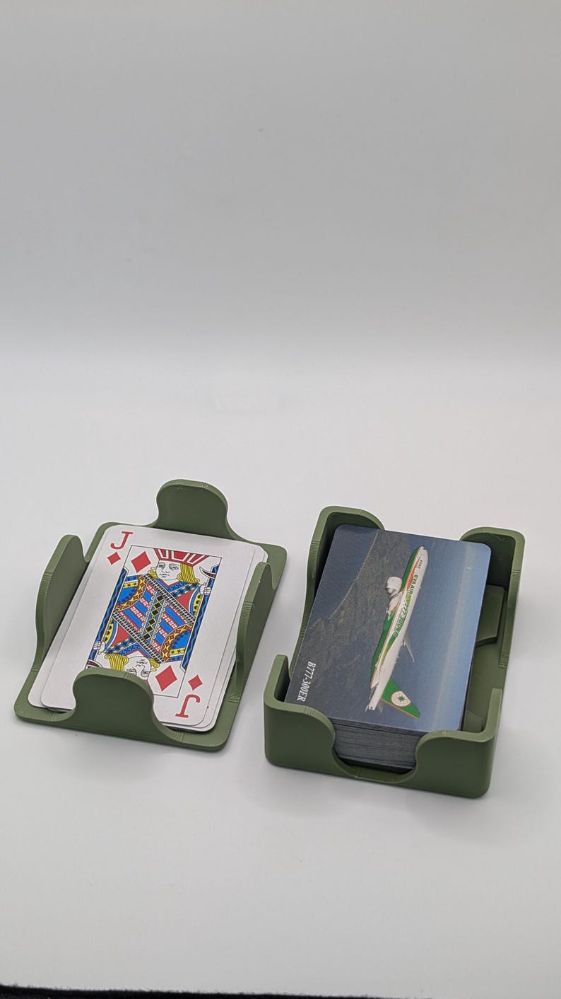 May include: Two olive green card holders, each containing playing cards. One holder displays a Jack of Diamonds card. The other holder contains cards with a green and white design. The holders are on a white surface.