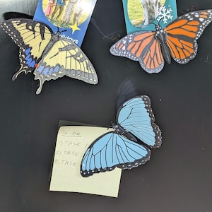 May include: A collection of butterfly-shaped magnets in various colors, including yellow, orange, and blue. The magnets are placed near small cards with images and text, including a family photo and a husky dog. A yellow sticky note with a "To Do" list is also present.