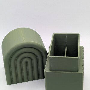 May include: Olive green 3D-printed desk organizer with a rectangular base and a divided interior. A matching arched lid sits beside it. The organizer has a modern, minimalist design.