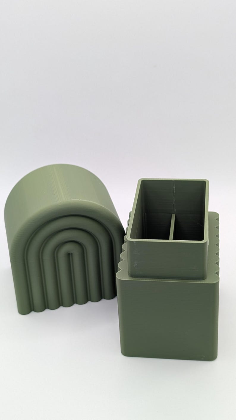 May include: Olive green 3D-printed desk organizer with a rectangular base and a divided interior. A matching arched lid sits beside it. The organizer has a modern, minimalist design.