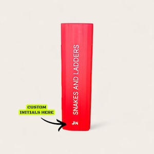 May include: A red board game box for Snakes and Ladders, with the game title printed vertically in white. The bottom left corner features a yellow box with the text "CUSTOM INITIALS HERE" and an arrow pointing to the game box.