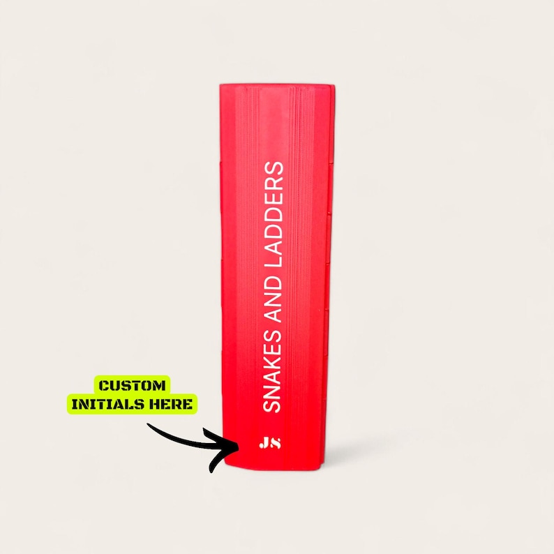 May include: A red board game box for Snakes and Ladders, with the game title printed vertically in white. The bottom left corner features a yellow box with the text "CUSTOM INITIALS HERE" and an arrow pointing to the game box.