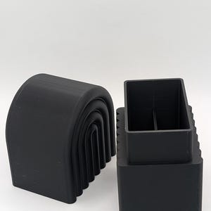 May include: Two black, 3D-printed objects. One is a curved, arch-shaped piece with a series of nested arches. The other is a rectangular box with a square opening and a divider inside. The objects are set against a white background.