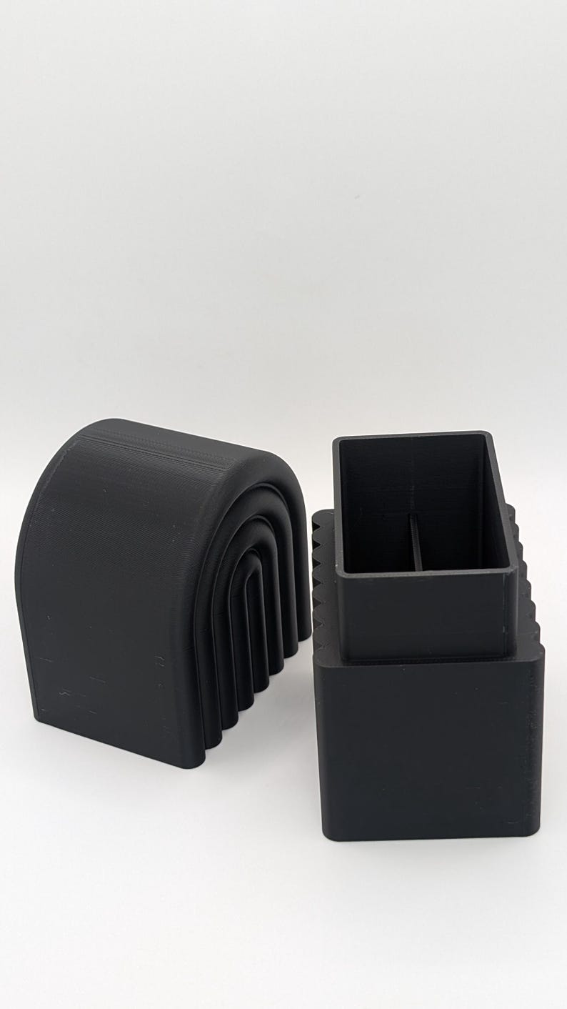May include: Two black, 3D-printed objects. One is a curved, arch-shaped piece with a series of nested arches. The other is a rectangular box with a square opening and a divider inside. The objects are set against a white background.