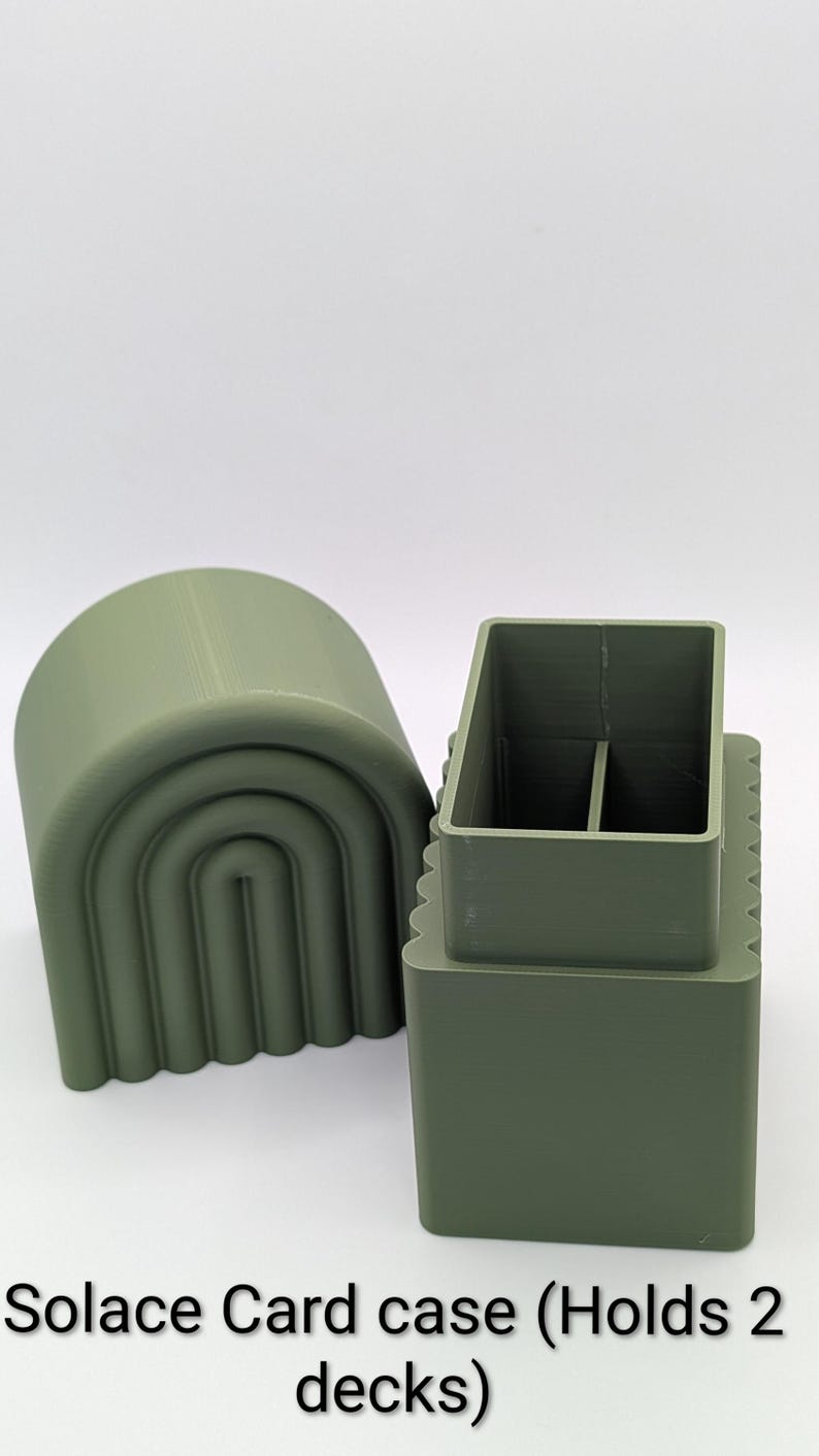 May include: A sage green card case with a matching arched lid. The case is rectangular with a divided interior, designed to hold two decks of cards. The text "Solace Card case (Holds 2 decks)" is printed at the bottom.