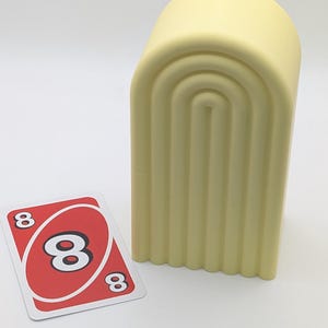 May include: A light yellow, arched container with vertical grooves, next to an Uno card. The card is red with a white center and three black number 8s. The background is plain white.