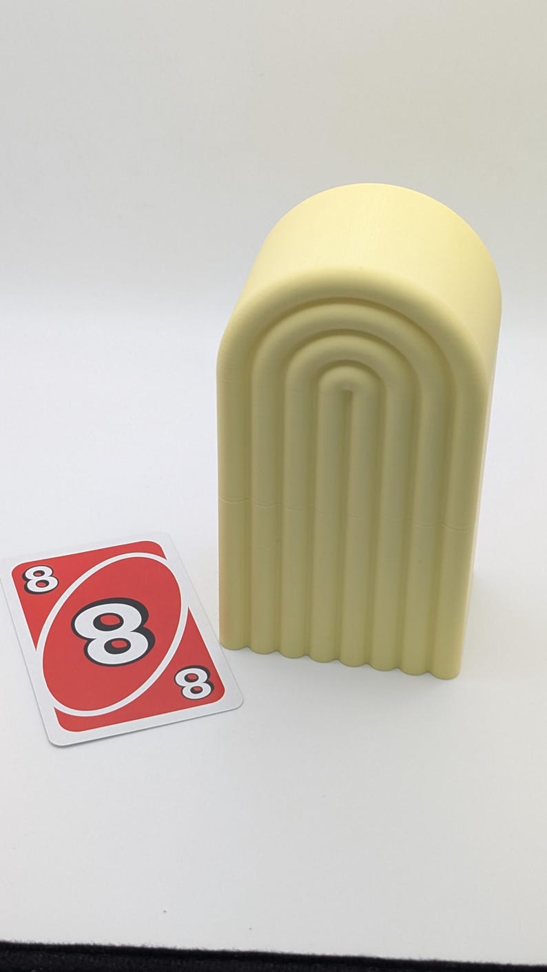 May include: A light yellow, arched container with vertical grooves, next to an Uno card. The card is red with a white center and three black number 8s. The background is plain white.