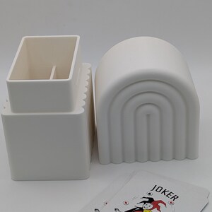 May include: A still life composition featuring playing cards and decorative objects. The cards display a colorful illustration of a joker. Two white ceramic containers are also present, one rectangular and one arched.