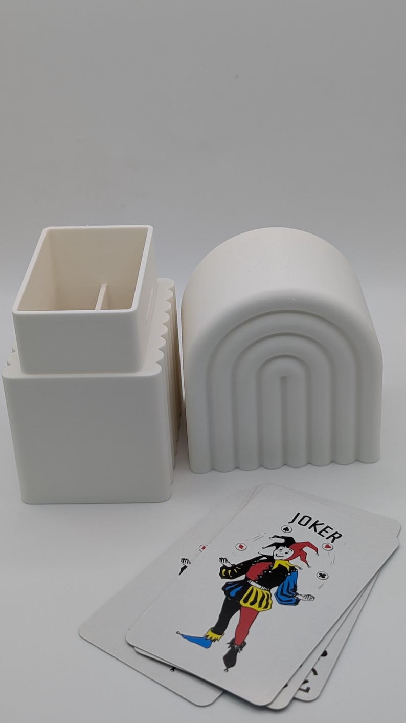 May include: A still life composition featuring playing cards and decorative objects. The cards display a colorful illustration of a joker. Two white ceramic containers are also present, one rectangular and one arched.