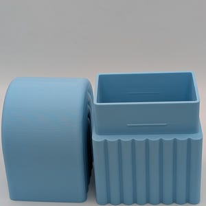 May include: Two light blue, 3D-printed objects. One is a cylindrical shape with a rounded top and bottom. The other is a rectangular box shape with a ridged base and a separate lid. Both objects are made of plastic.