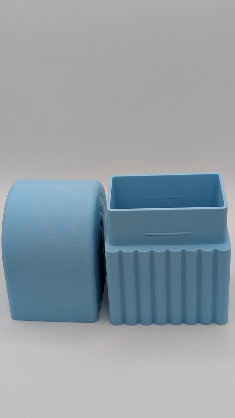 May include: Two light blue, 3D-printed objects. One is a cylindrical shape with a rounded top and bottom. The other is a rectangular box shape with a ridged base and a separate lid. Both objects are made of plastic.