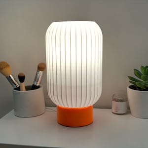 May include: A white, ribbed table lamp with an orange base is illuminated. Makeup brushes in a gray holder, a small potted succulent, and a candle sit nearby. The lamp is the focal point.