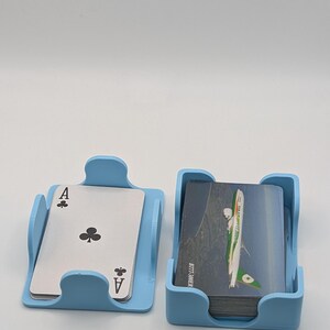May include: Two light blue card holders, one with an Ace of Clubs playing card and the other with a stack of cards. The cards in the second holder have a green and white airplane design. The holders are made of plastic.