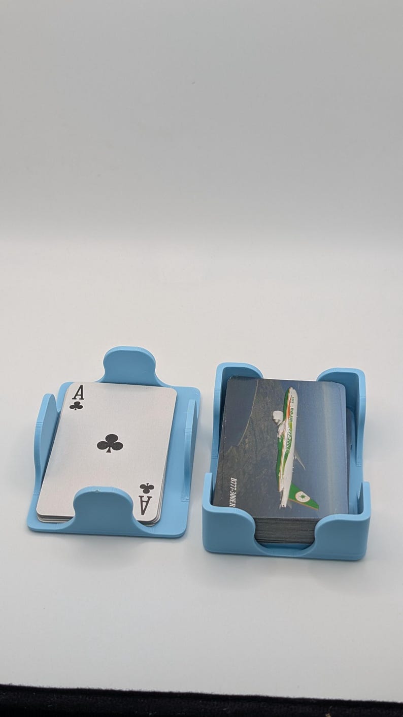 May include: Two light blue card holders, one with an Ace of Clubs playing card and the other with a stack of cards. The cards in the second holder have a green and white airplane design. The holders are made of plastic.