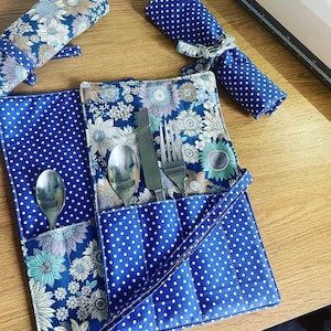 Op de afbeelding: Een blauwe en witte stoffen bestekrol met een bloemenpatroon en witte stippen. De rol heeft een band en bevat een vork, een mes en twee lepels.