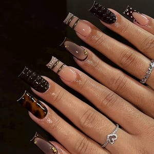 May include: Long square-shaped artificial nails featuring a mix of designs including solid dark brown, tortoiseshell pattern, and French tips with gold metallic accents, rhinestones, and cable-knit textures, worn with two silver-toned rings.